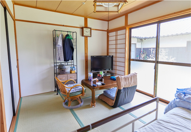 和室の居室。畳敷きで木造建築。テレビ、ラタン椅子、衣類掛け、ベッドが見える。